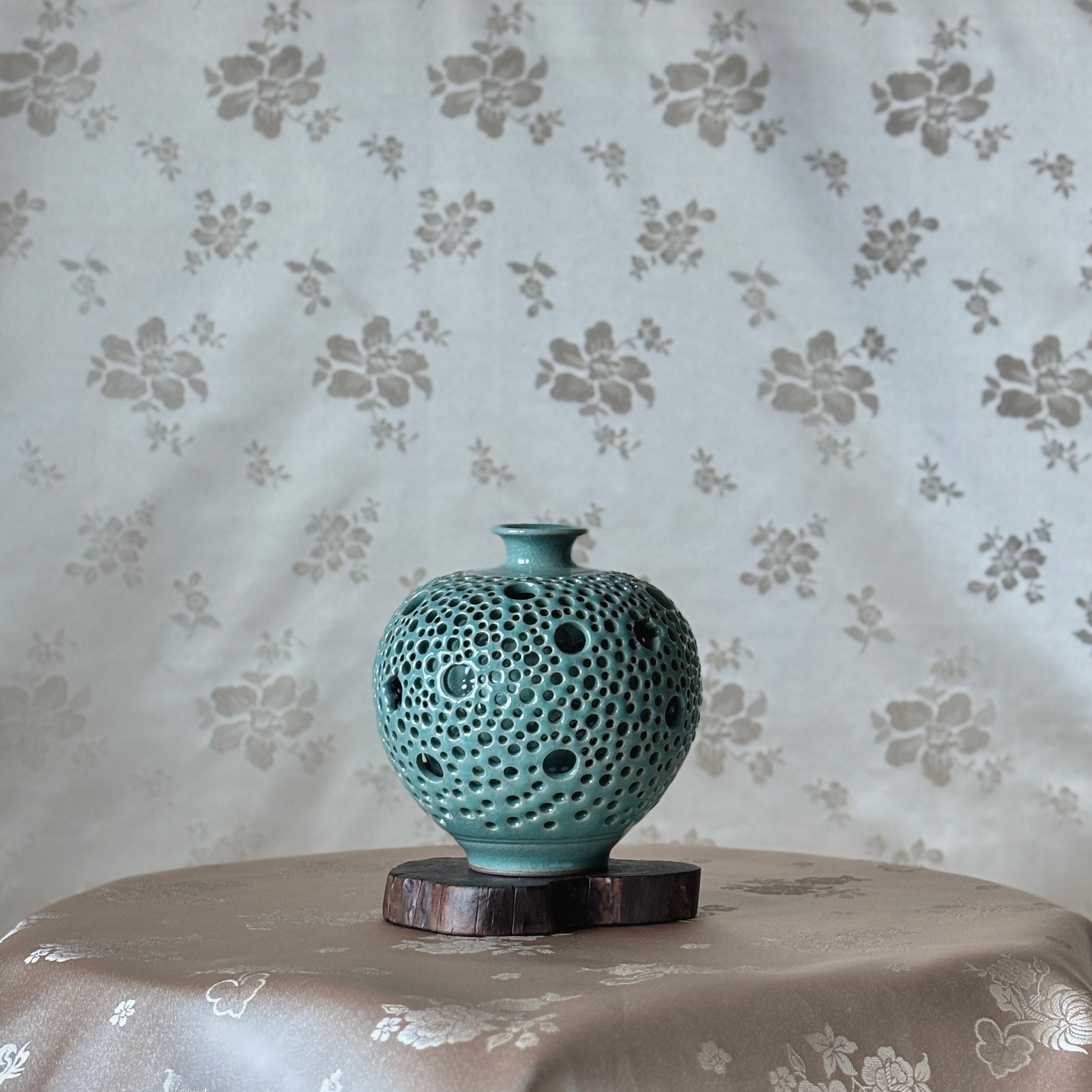 korean ceramic vase with textured surface on a wooden stand against floral-patterned wallpaper.