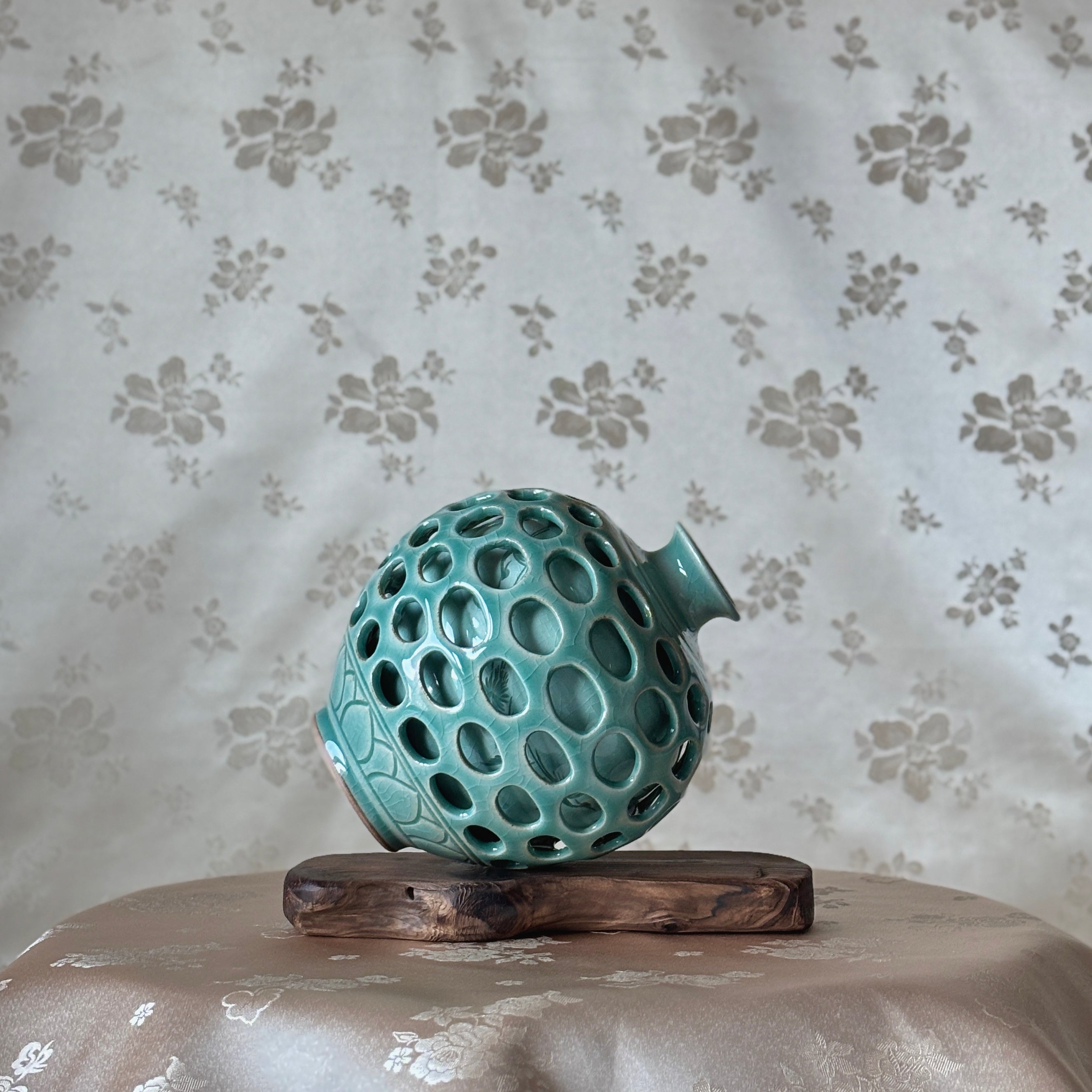 celadon vase with a unique textured design on a wooden stand against a floral-patterned wall.