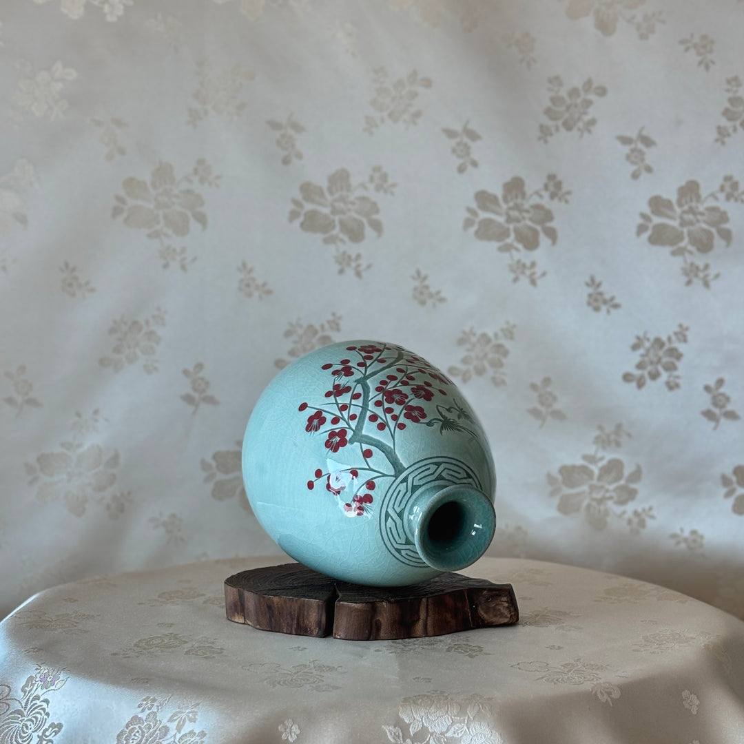 Light green ceramic vase with floral design on a wooden stand against a floral wallpapered wall.